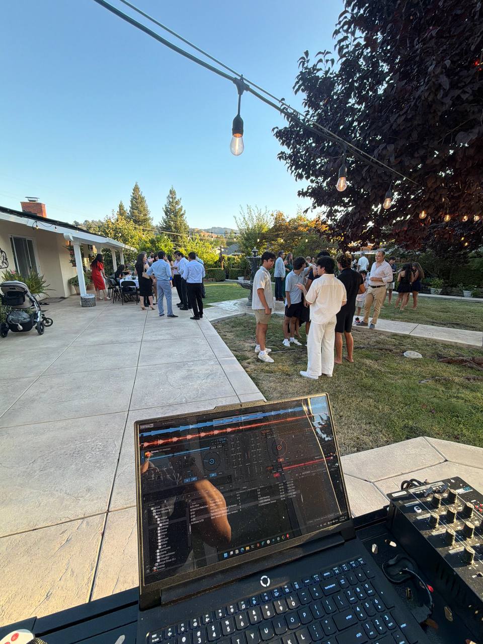 Guests gathered around DJ Alphire's laptop and controller at golden hour under string lights