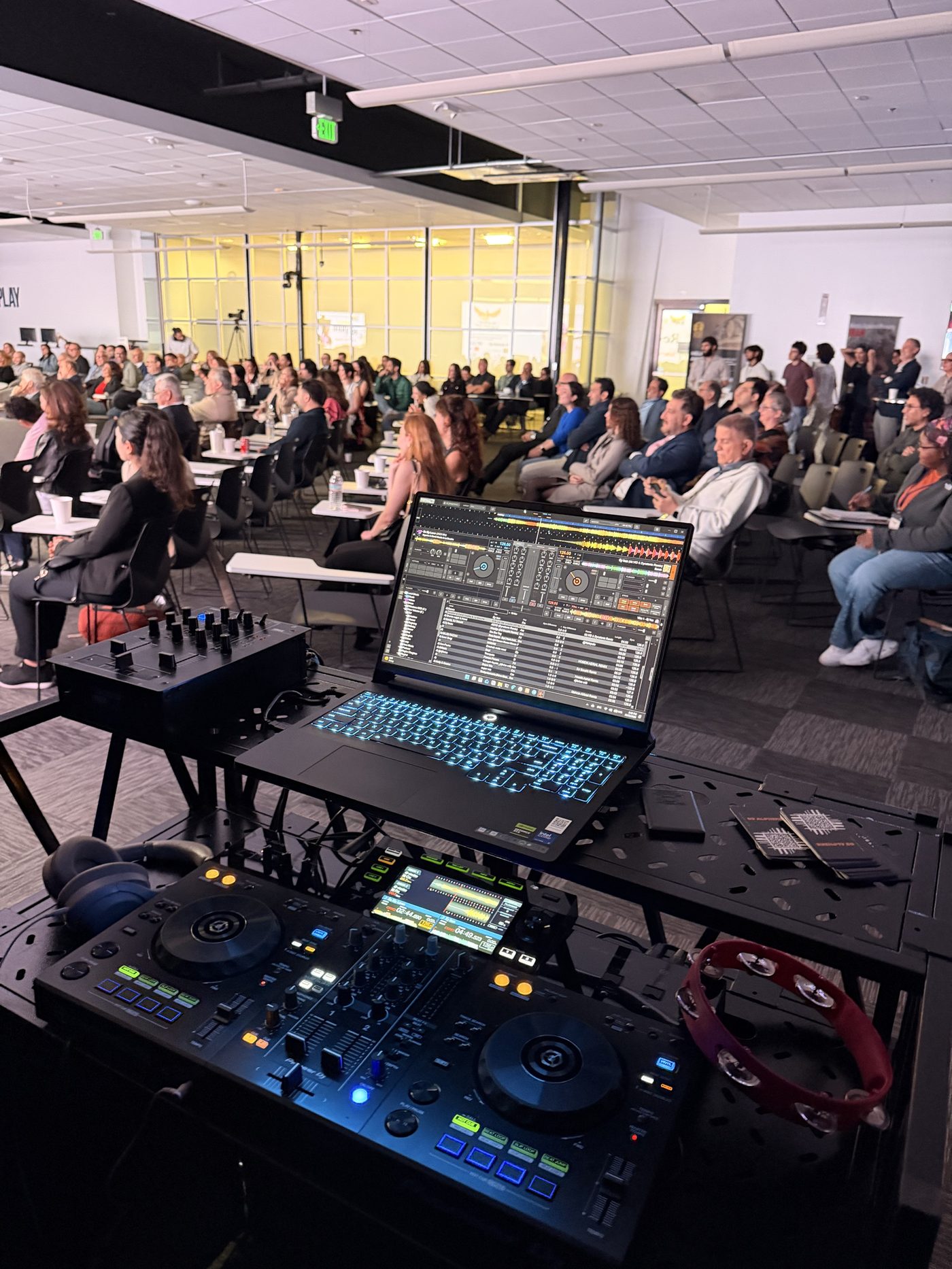 DJ Alphire's Pioneer DJ setup and laptop facing a full seated audience at Plug and Play Sunnyvale -- October 2026 corporate event